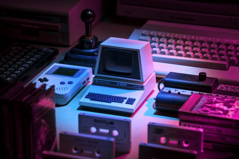 Retro computers, a handheld game console, and cassette tapes under pink light